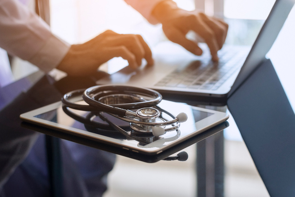 A desktop with a stethoscope sitting on a tablet and a physician typing on a laptop